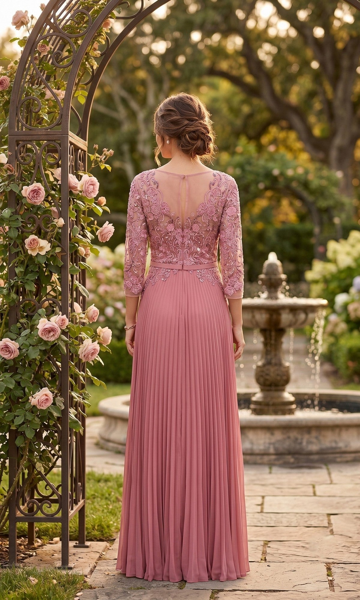 Wearing the Long Formal Dress: Chicas YG5038, a person stands with their back to the camera in a garden by a fountain and blooming roses. The dress features mauve lace, pleats, and an embroidered bodice for a modest formal look.